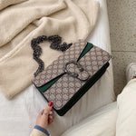 Patterned handbag with chain strap held by a hand on a soft surface.