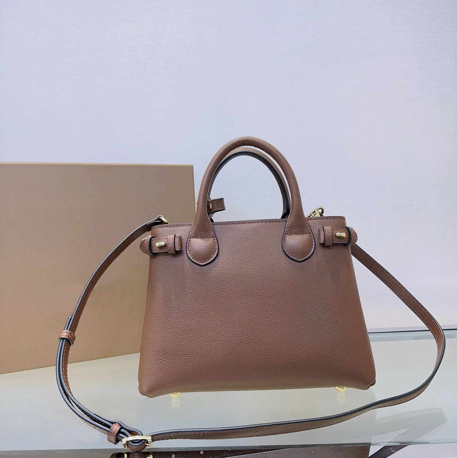 Brown handbag with a matching strap on a clear surface with a beige box in the background.