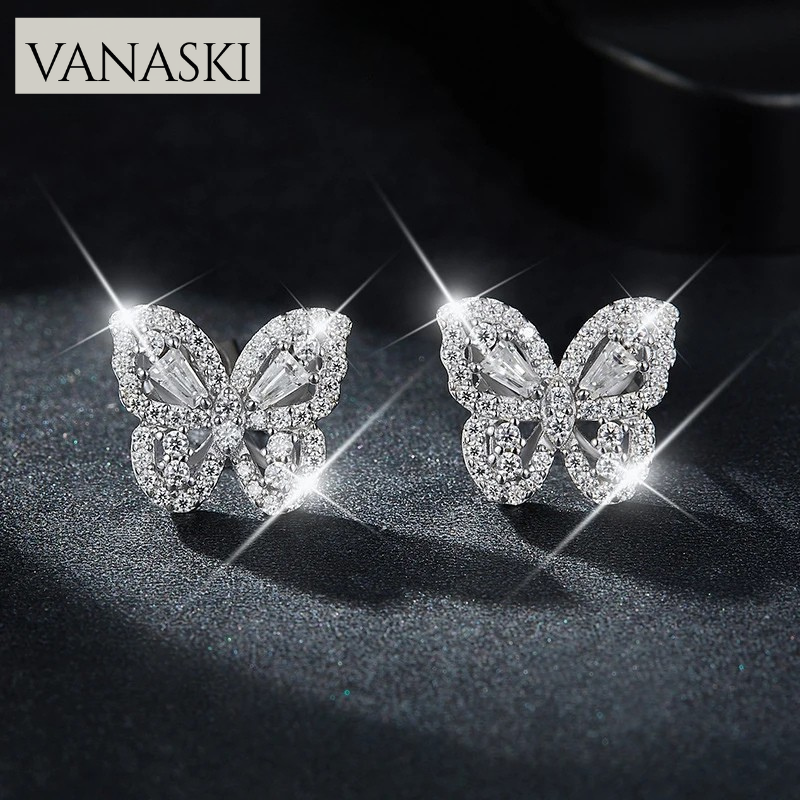 Butterfly-shaped earrings with sparkling stones on a dark background, featuring the VANASKI brand.