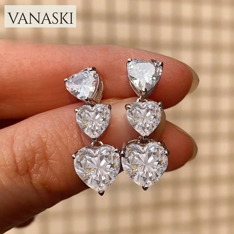 Heart-shaped earrings held between fingers with 'VANASKI' brand name above.