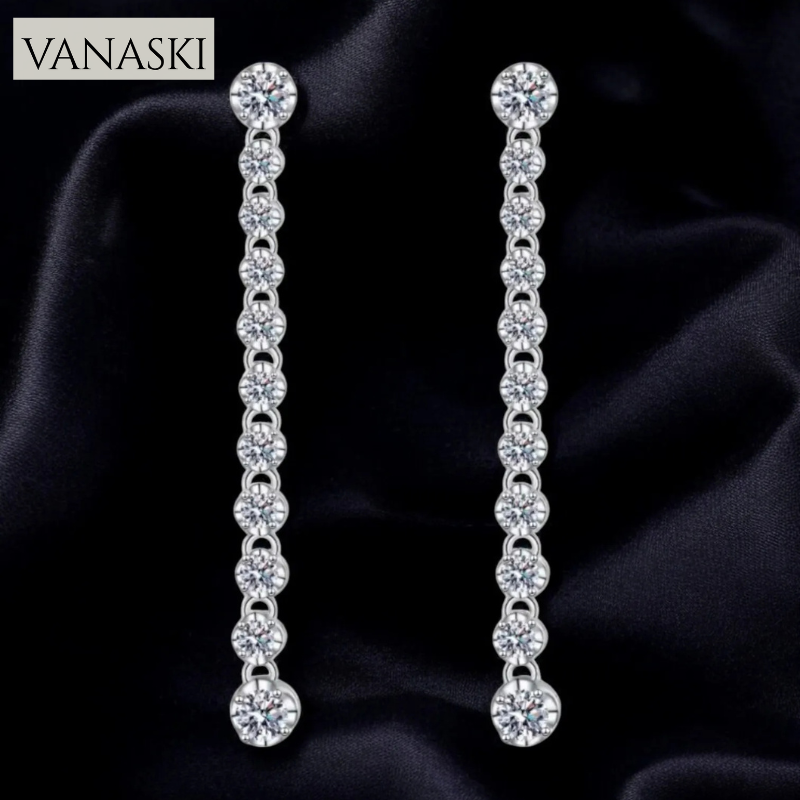 Pair of silver earrings with clear stones on a dark fabric background, branded 'VANASKI'.