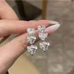 Heart-shaped diamond earrings held by a hand against a blurred background