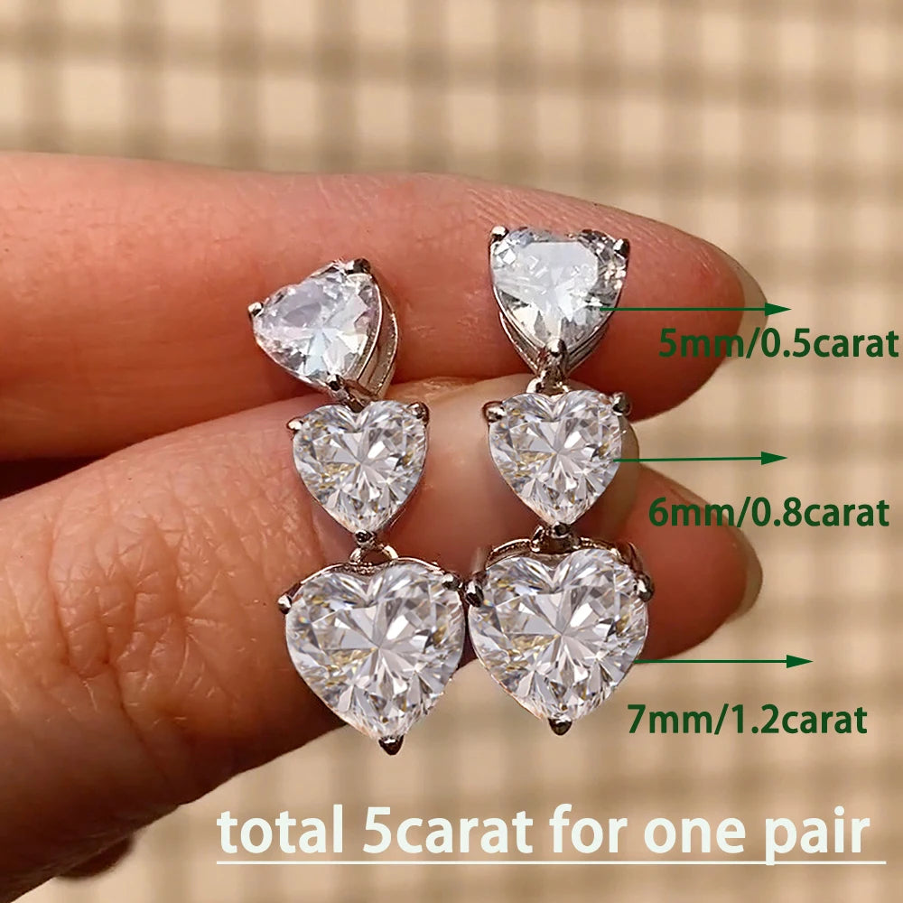 Heart-shaped diamond earrings held between fingers with size labels on a blurred background