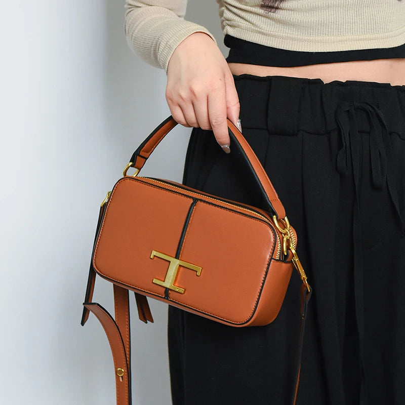 Brown handbag with gold 'T' logo held by a person wearing black pants and a beige top.