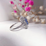 Silver ring with a blue gemstone on a white surface with blurred flowers in the background