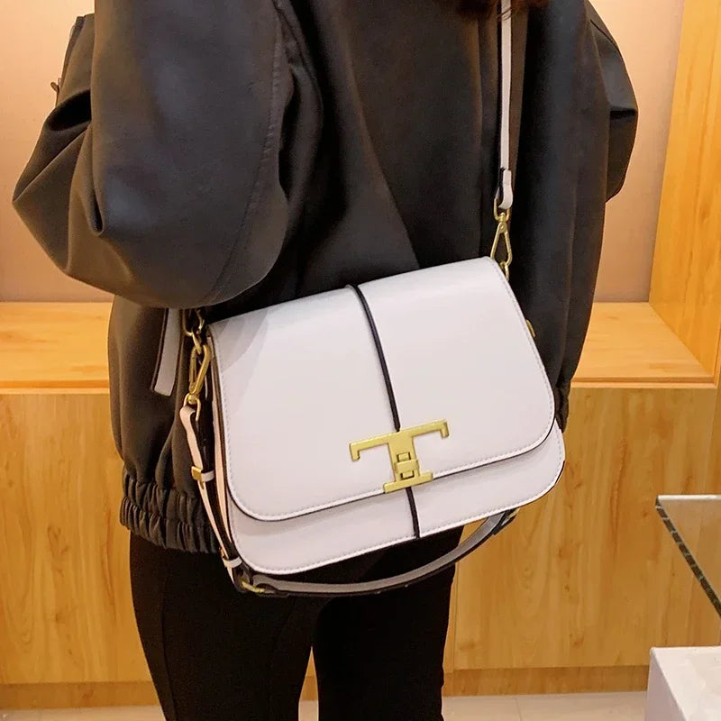 White handbag with gold 'T' logo worn by a person against a wooden background