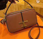Brown handbag with gold 'T' logo held by a hand on an orange surface.
