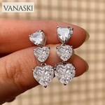 Heart-shaped earrings held between fingers with 'VANASKI' brand name above.