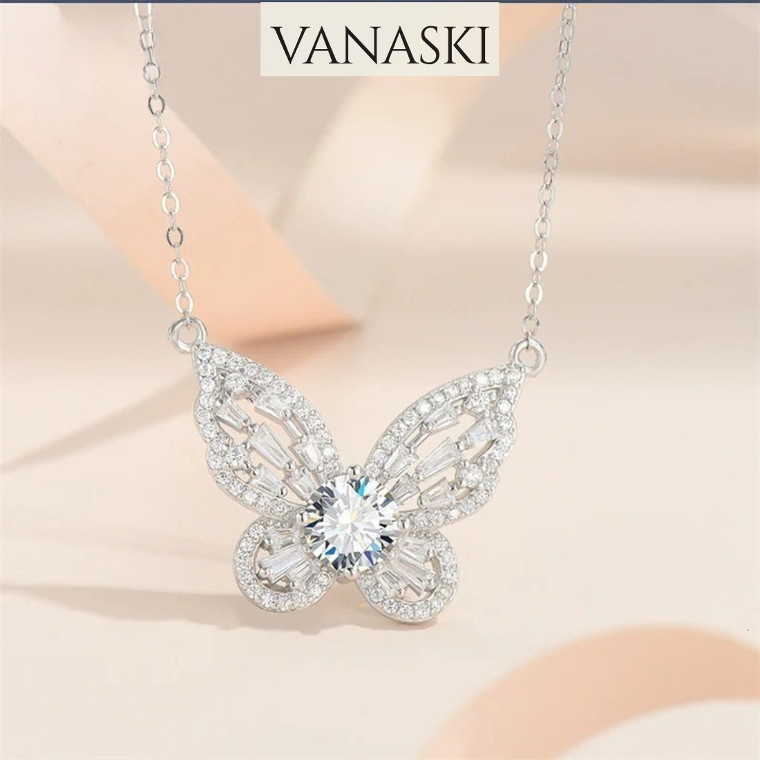 Butterfly-shaped necklace with sparkling stones on a soft beige background, featuring the brand 'VANASKI'.